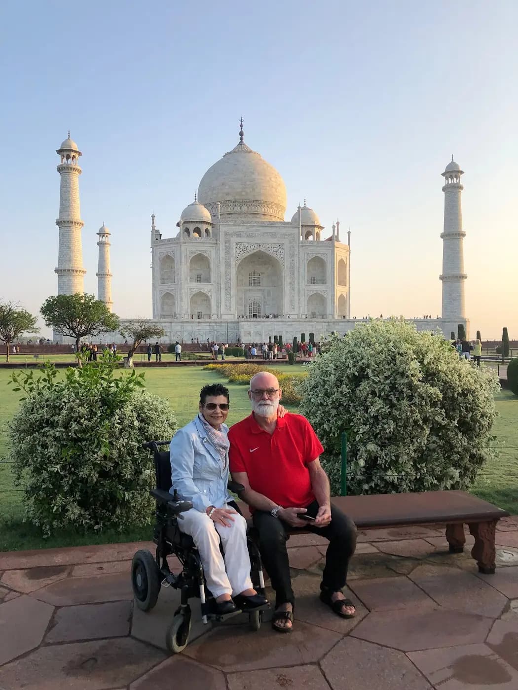 Una pareja de viajeros, ella en silla de ruedas, sentada junto a un banco en los jardines del Taj Mahal, en la India.